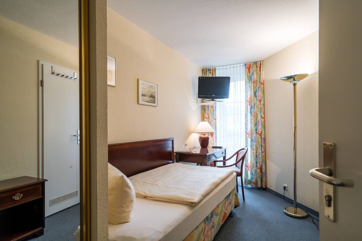 Quarto de hotel com uma cama, uma escrivaninha com luminária, televisão e janela com cortinas.