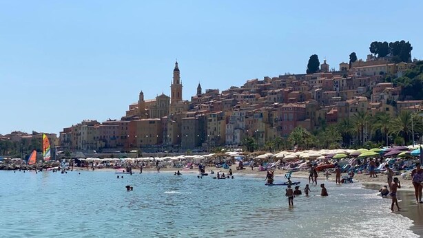 A beach with people swimming and sunbathing, a clear sky, and buildings in the background.