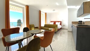 A hotel room with a bed, a dining table with chairs, a kitchen area, and a view of the sea.
