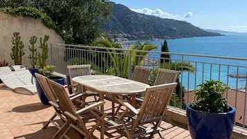 An outdoor seating area with a table and chairs, surrounded by potted plants and a view of the sea and mountains.