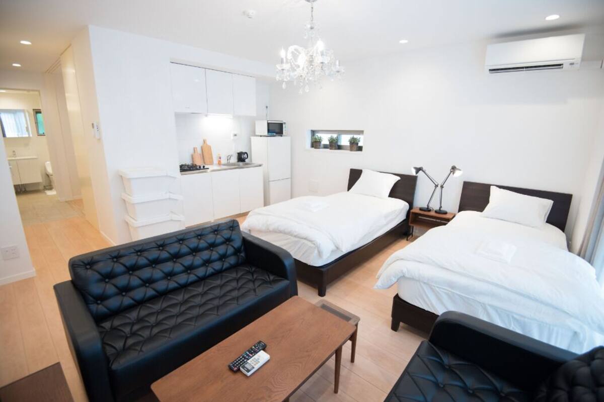 A modern hotel room with a sofa, two beds, a small kitchen area, and a chandelier.