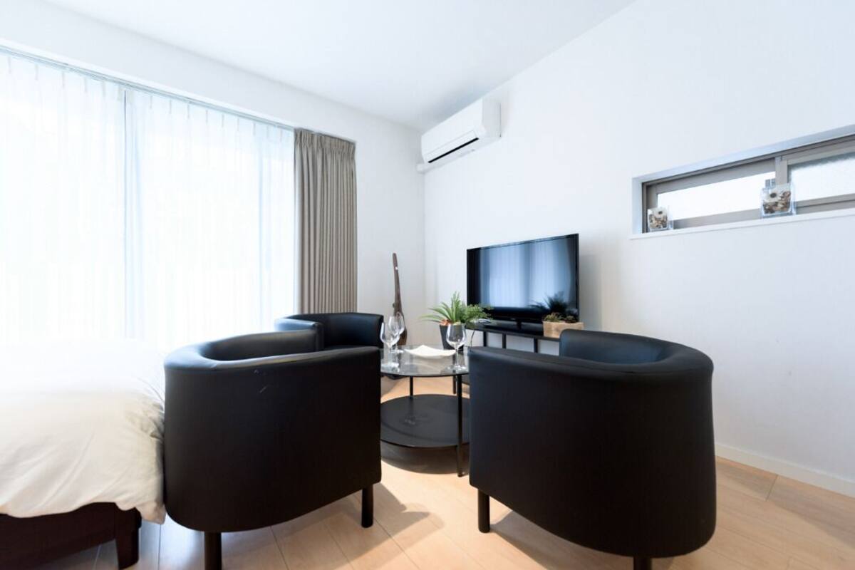 A modern living room with a bed, two black armchairs, a small round table, a TV, and a window with curtains.