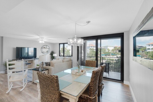 A modern living room with a glass dining table, wicker chairs, a flat-screen TV, and a balcony with a view of the neighborhood.