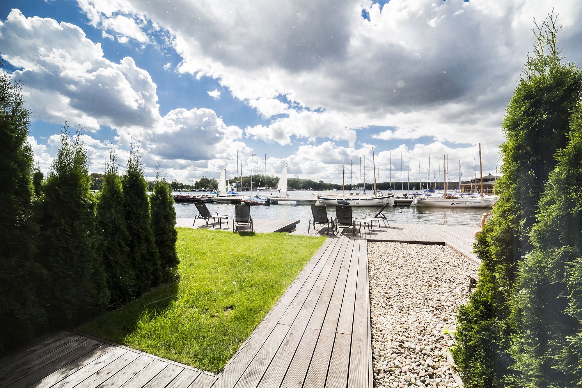 Eine Holzterrasse führt zu einem Yachthafen mit dort liegenden Booten, umgeben von viel Grün und einem bewölkten Himmel.