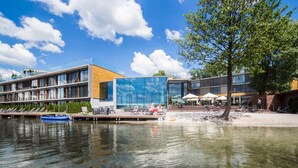 A modern building with a glass facade, a wooden deck with lounge chairs, and a dock with a boat. - Przystań Hotel & Restaurants (Olsztyn)