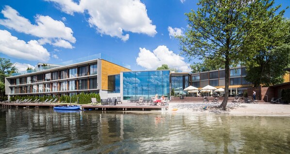 Un edificio moderno con fachada de vidrio, una terraza de madera con sillones de descanso y un muelle con un bote.