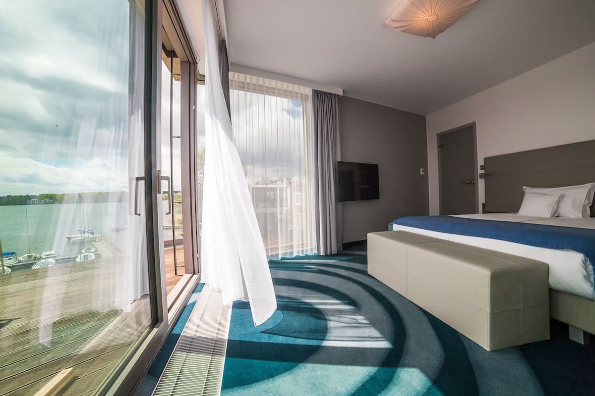 A modern hotel room with a large window, a bed, a TV, and a view of the water.