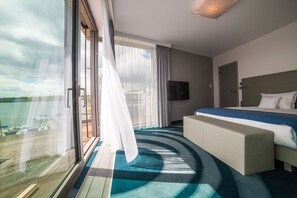 A modern hotel room with a large window, a bed, a TV, and a view of the water.
