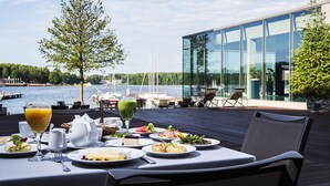 A dining table with breakfast items, overlooking a lake with boats and a modern building. - Przystań Hotel & Restaurants (Olsztyn)