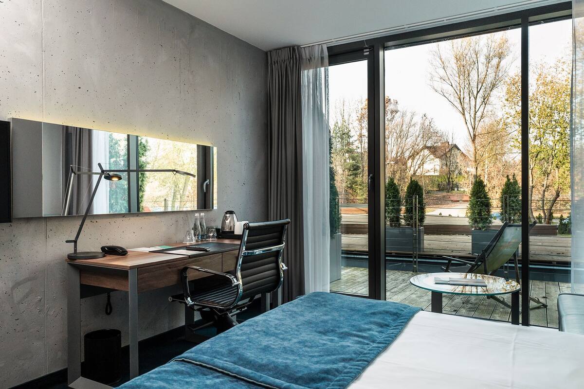 A modern hotel room with a large bed, a desk with a chair, a small table, and a view of the outdoors.