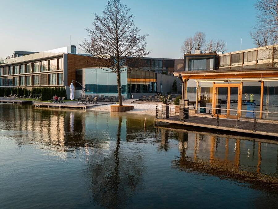 Un edificio moderno con fachada de vidrio, terraza de madera y reflejo en el agua.