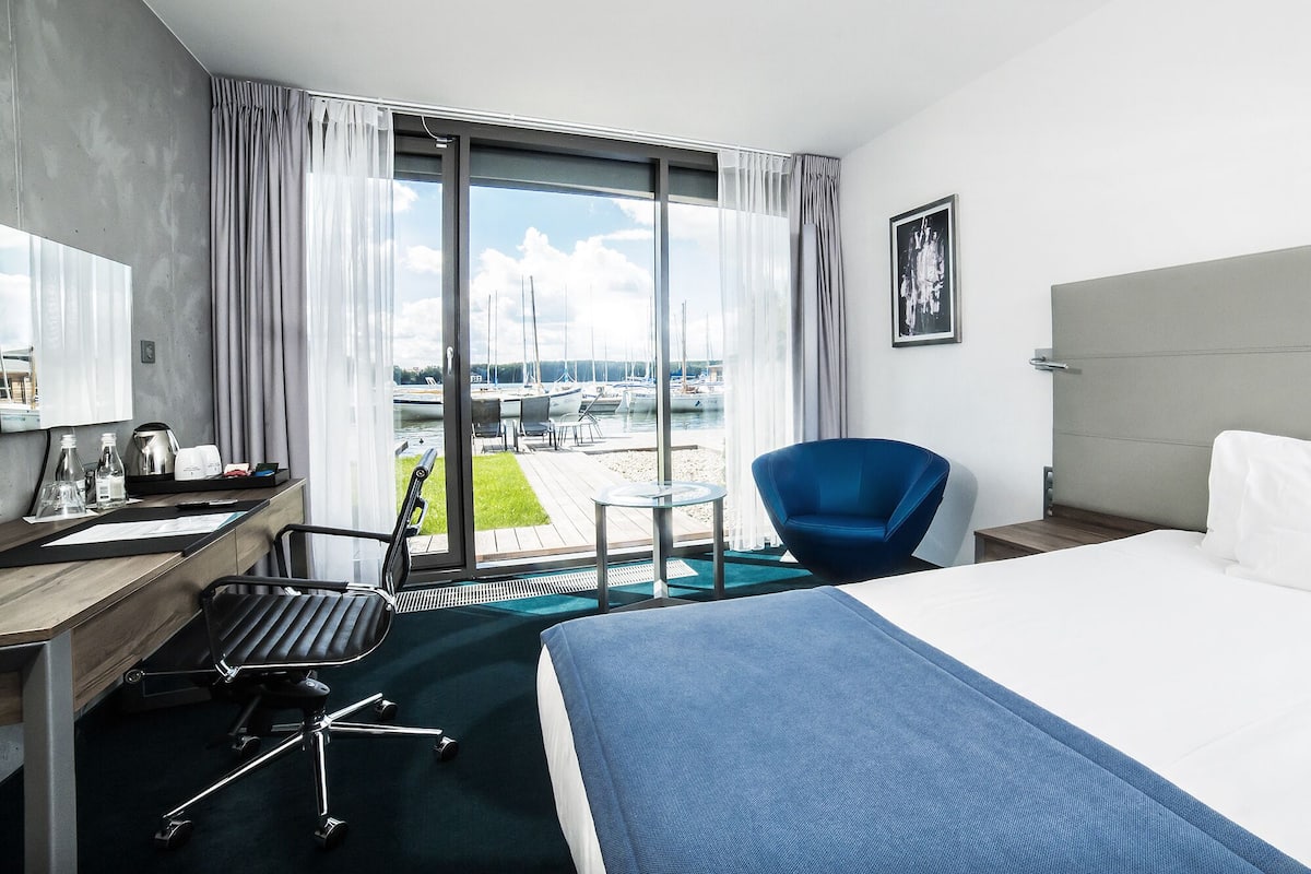 A hotel room with a large bed, a desk with a chair, a small table, and a view of a marina with boats.