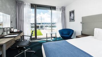 A hotel room with a large bed, a desk with a chair, a small table, and a view of a marina with boats.
