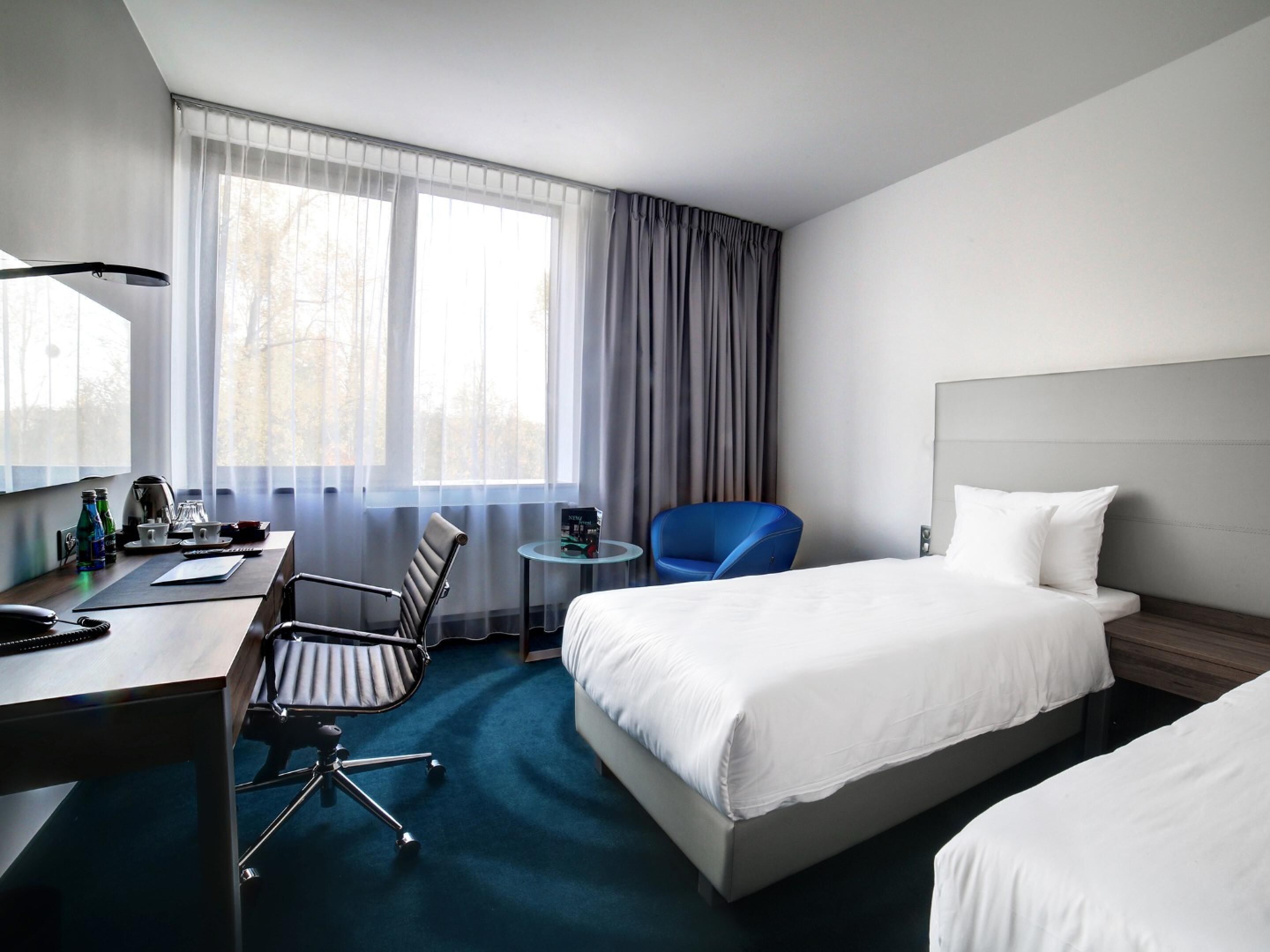 A hotel room with a desk, two chairs, a bed, and a window with curtains.