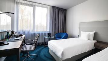 A hotel room with a desk, two chairs, a bed, and a window with curtains.