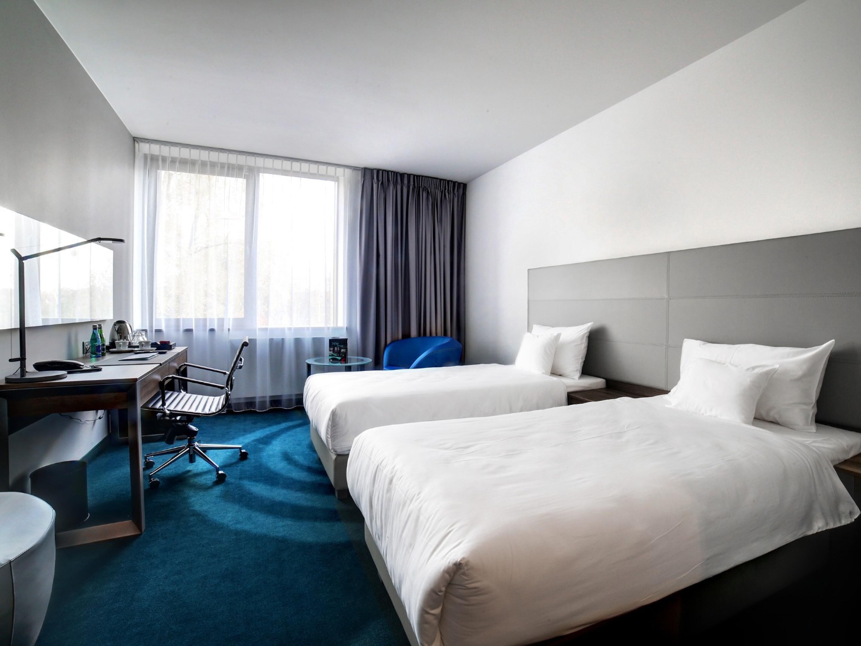 Habitació d'hotel amb dos llits, una taula de treball, una cadira, una làmpada i una finestra amb cortines.