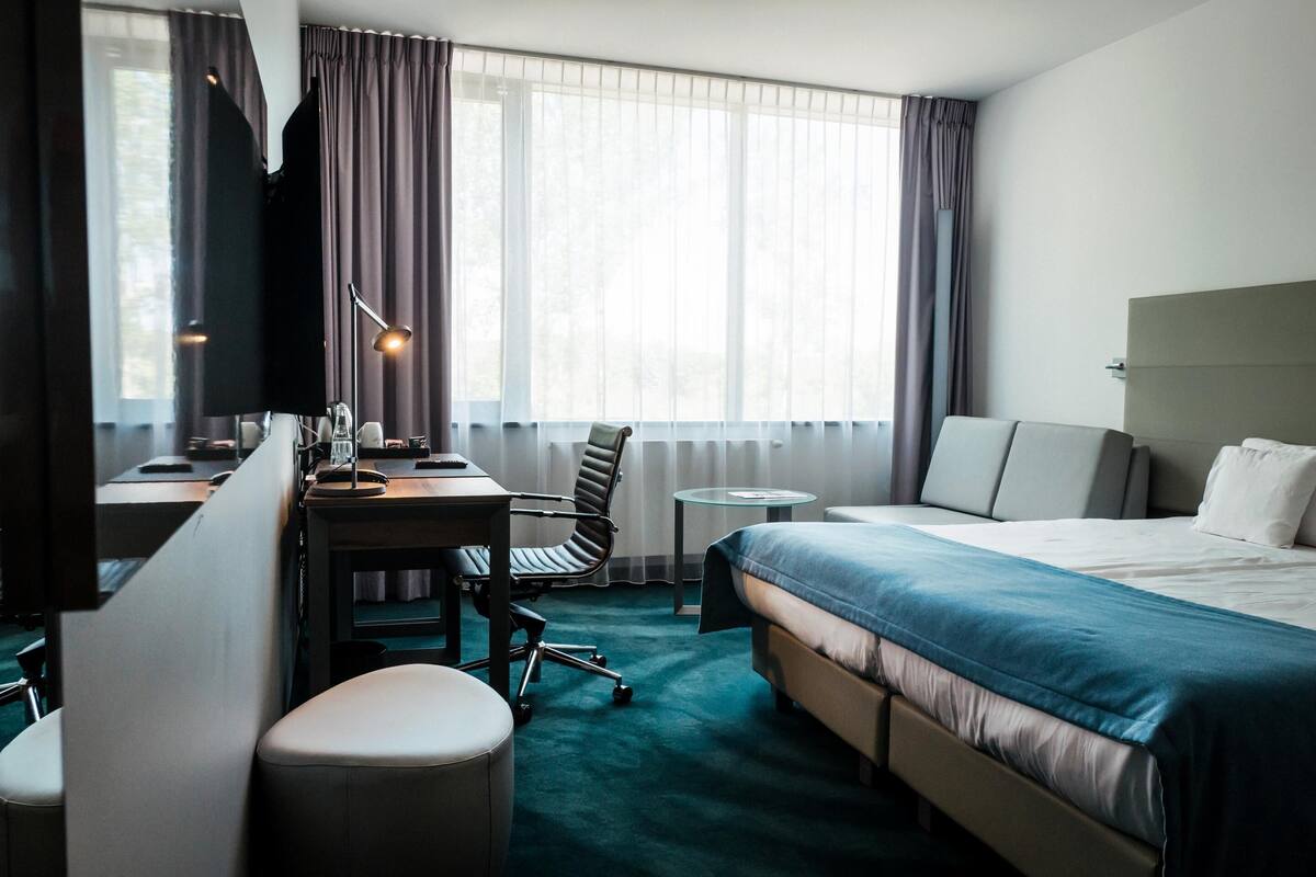 A hotel room with a bed, a desk, a chair, a small table, and a window with curtains.