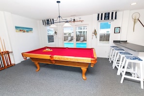Una mesa de billar en una habitación con vista a una piscina exterior, una zona de bar con taburetes e iluminación en el techo.