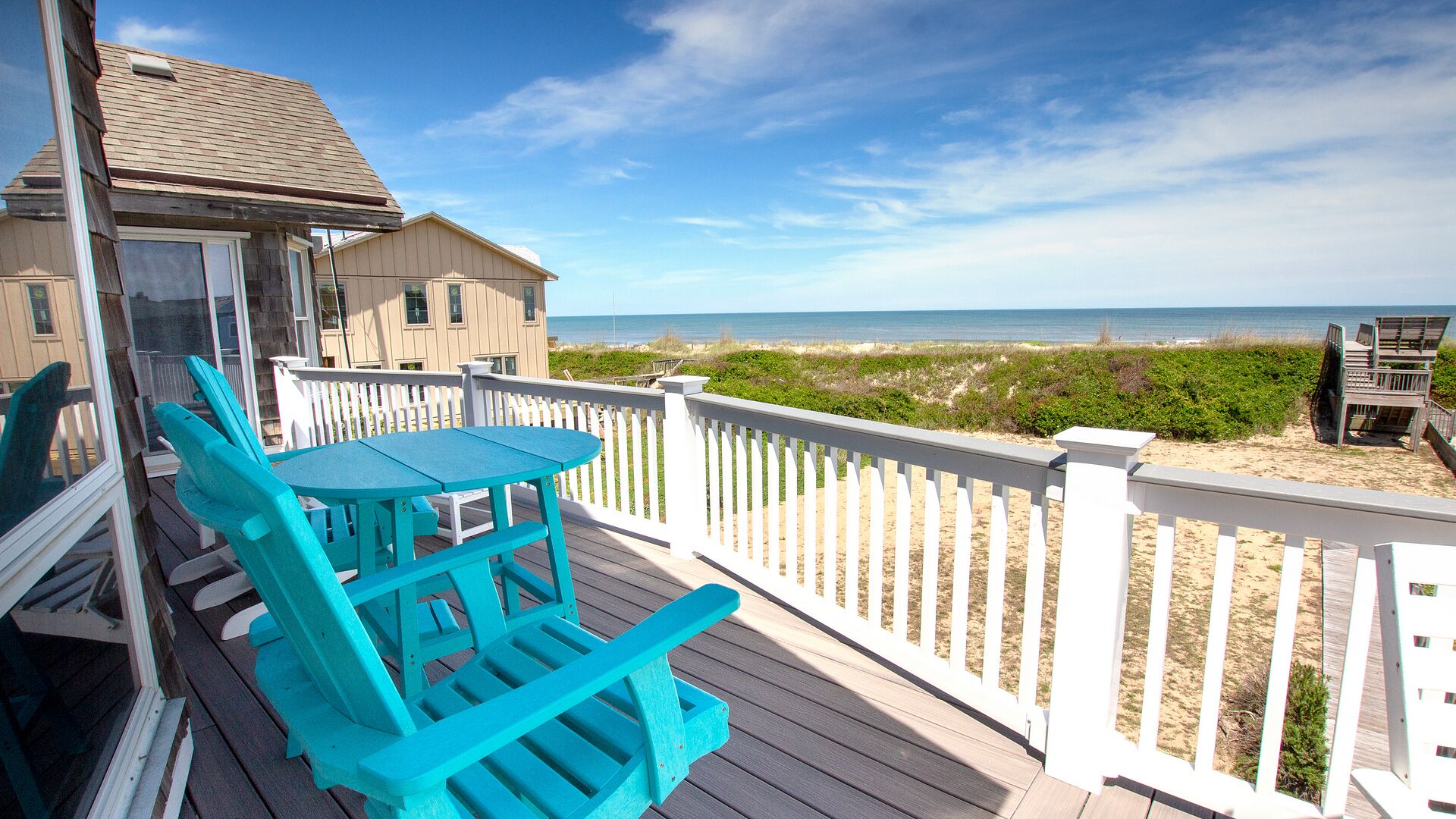 A wooden deck with a table and chairs, overlooking a beach with a white fence and a building in the background.