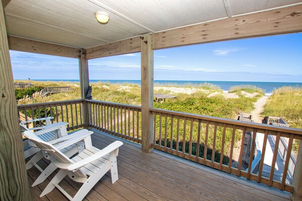 Una terraza de madera con sillas Adirondack blancas, vista a la playa y un sendero arenoso que lleva al océano.