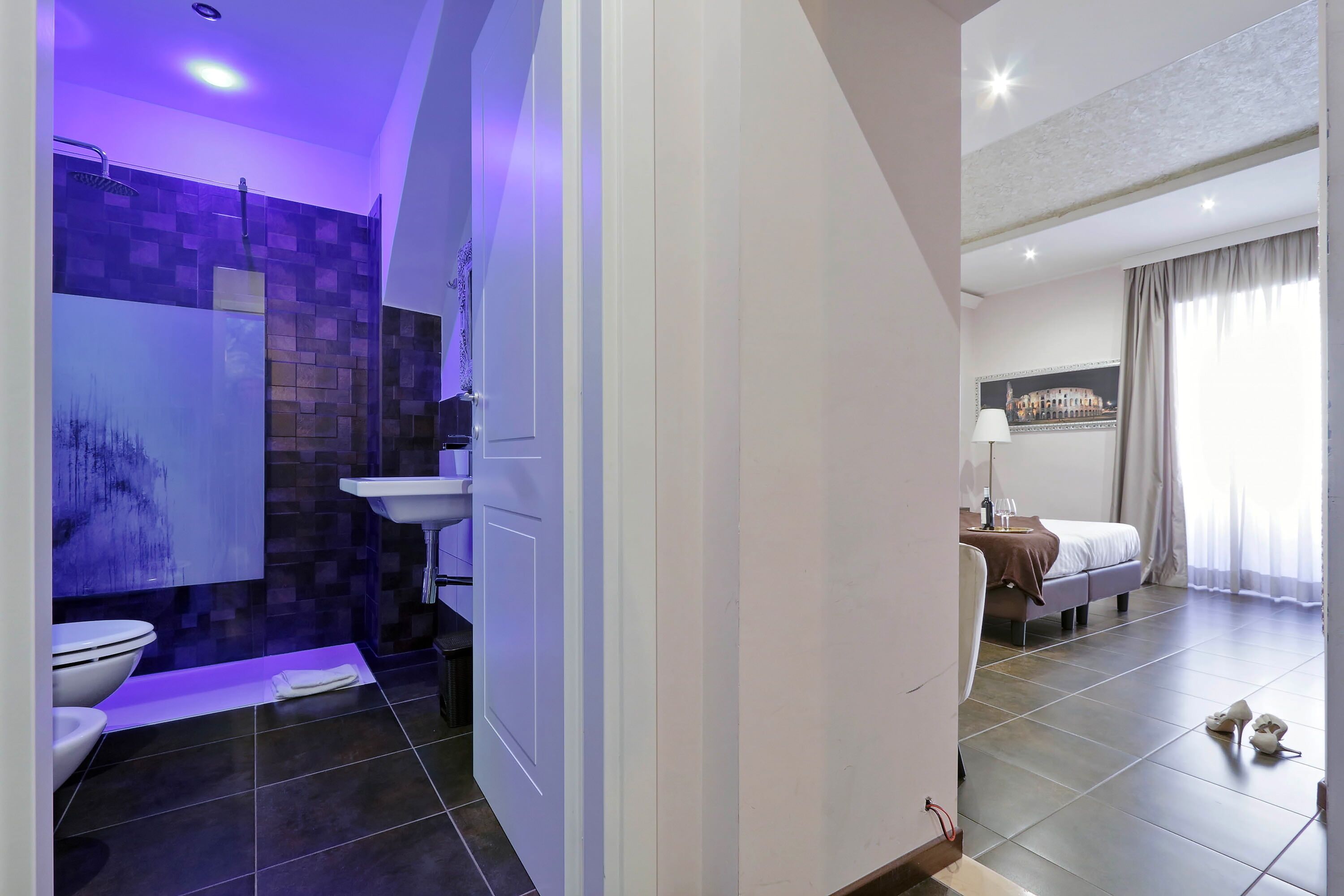 A modern bathroom with a shower, sink, and toilet. There is a bed with a nightstand and a lamp in the adjacent bedroom.
