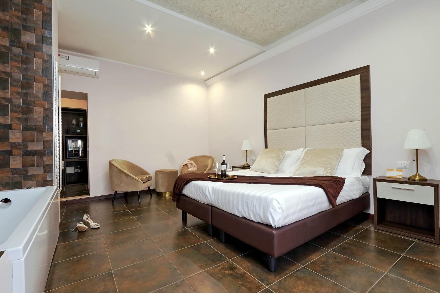 A hotel room with a large bed, a sitting area with two chairs, a bedside table with a lamp, and a minibar.
