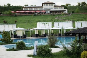 A pool area with white canopies and lounge chairs, a building with multiple windows in the background, and a grassy hill.