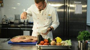 Een kok in een witte uniform met rode knopen, die een witte kom vasthoudt, staat in een keuken met roestvrijstalen apparatuur en diverse verse groenten op het aanrecht.