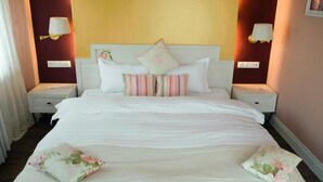 Een net opgemaakt bed met witte lakens en meerdere kussens, waaronder gestreepte en bloemenmotieven, in een kamer met gele muren en twee bedlampjes.