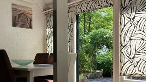 A room with a dining table, chairs, a white brick wall, a wooden ceiling, and a view of a garden through patterned curtains. - 5 Pers - Equipped House Gudrun With Sauna - Dogs are Welcome (Anjum)