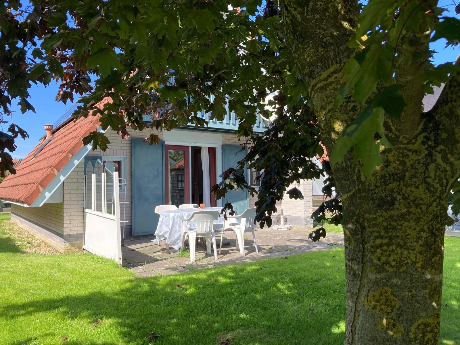 A house with a sloped roof, a covered patio with a table and chairs, and a tree in the foreground.