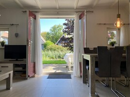 A room with a television, a dining table with chairs, and a view of a garden and house through open curtains.