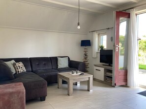 A living room with a sofa, a coffee table, a lamp, a television, and a door leading to an outdoor area. - Charming Bungalow Rental in Anjum With River Views (Anjum)