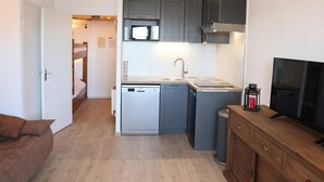 A compact kitchen with grey cabinets, a microwave, a dishwasher, and a small sink. There's a wooden cabinet with a television on top. - Charming studio for 4 in the heart of Les Orres 1650 (Les Orres)