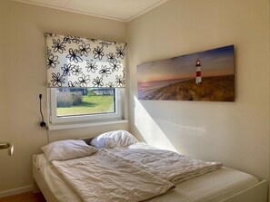 Un dormitorio con una cama, un ventanal con una persiana estampada de flores y un cuadro de un faro en la pared.
