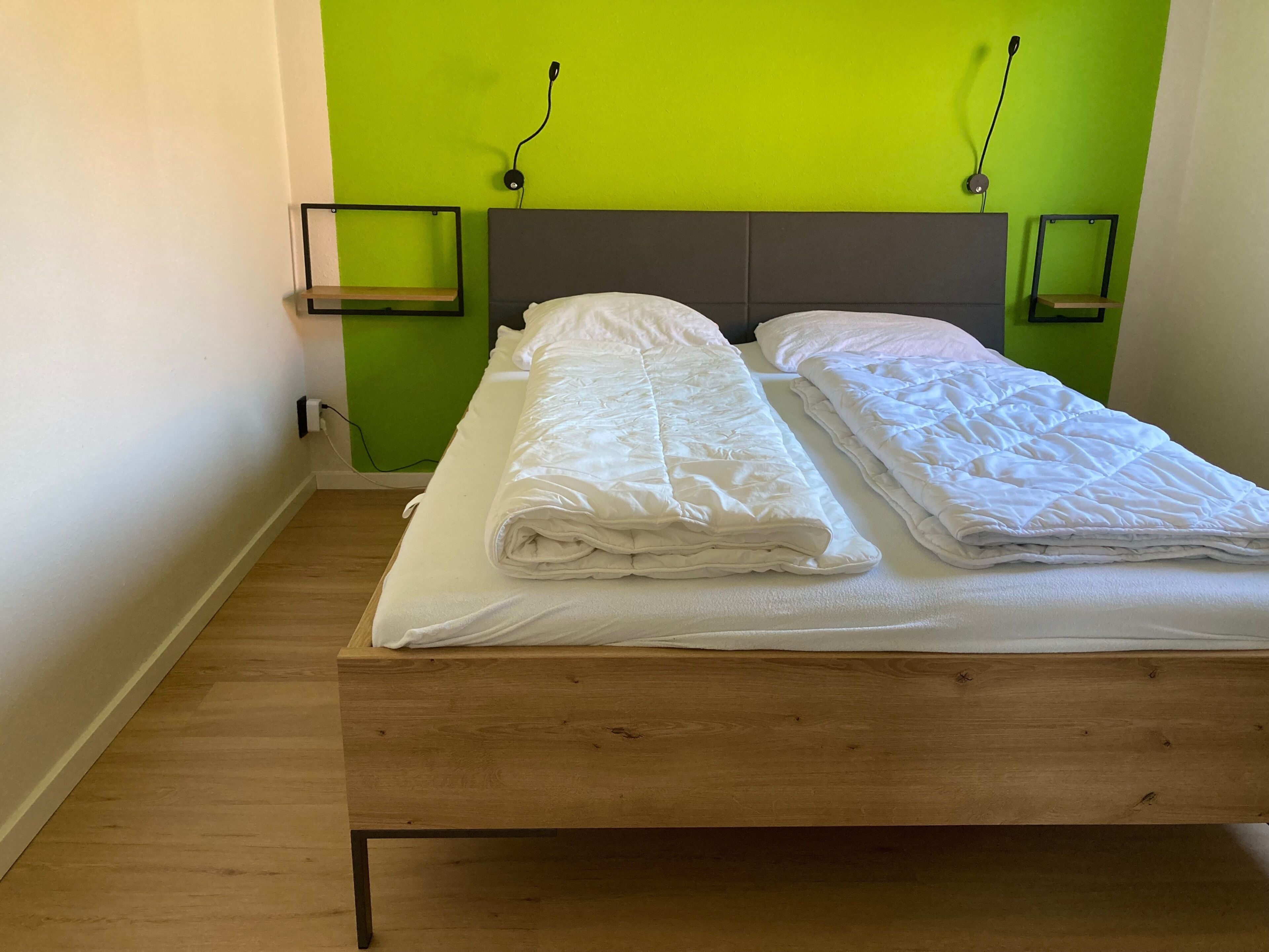 A single bed with a wooden frame and a grey headboard, positioned against a green wall with two black wall-mounted lamps.