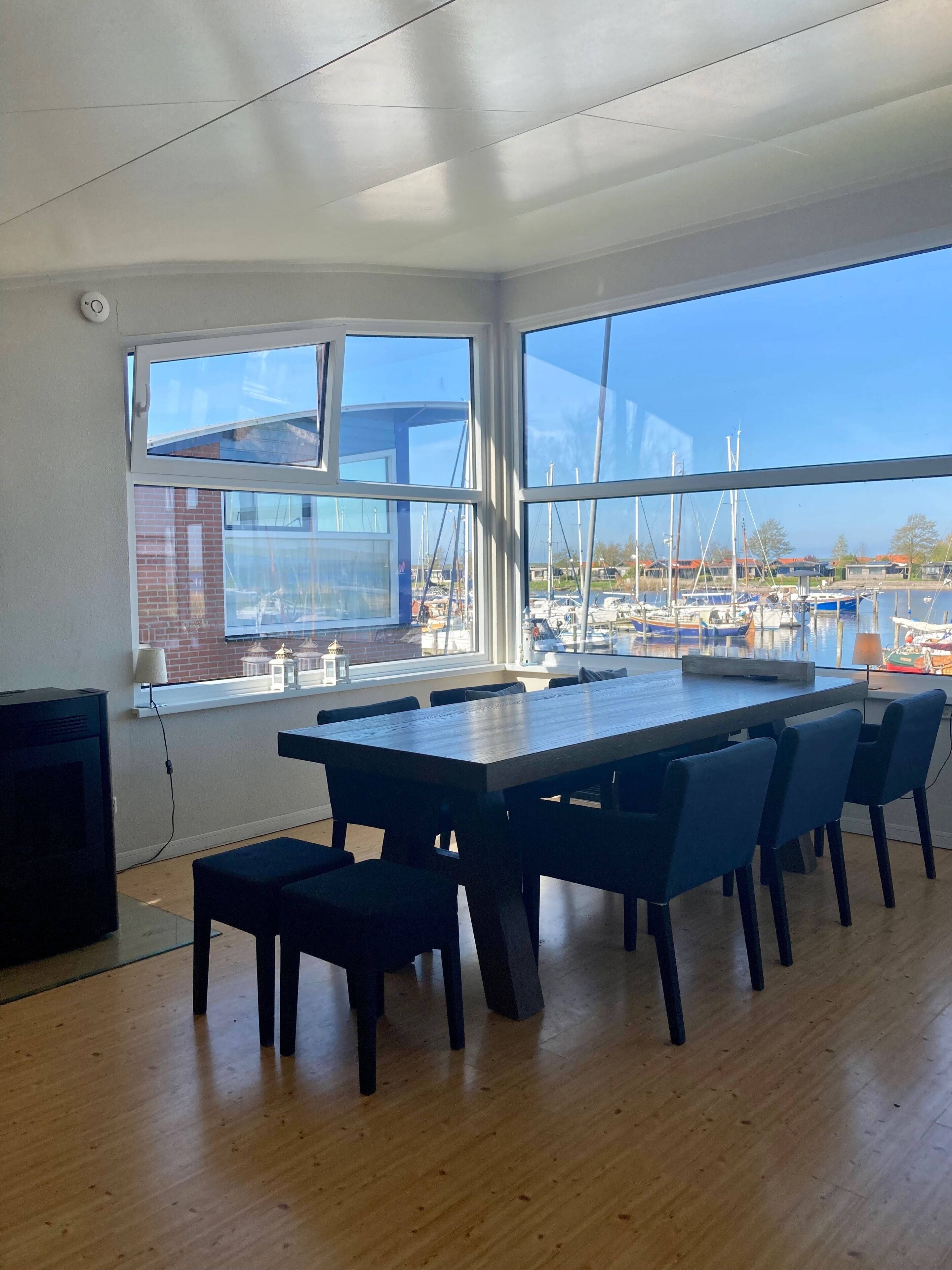 A spacious room with a large table and chairs, large windows with a view of boats and a building, and a TV on the left.