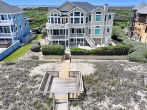 A multi-story beachfront property with a wooden deck leading to the entrance, surrounded by well-maintained hedges and a sandy landscape. - OCEANFRONT, Ocean Views, Pool, Rec Rm, PI244, Ashcroft (Corolla)