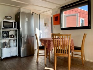 A compact kitchen with a refrigerator, microwave, and coffee maker. There is a dining table with chairs and a window with a view of a red building. - Chalet Gaby With Sauna, Private Dock, and Canoes at Lauwersmeer (Anjum)