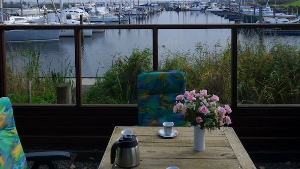 A wooden table with a kettle, cups, and a vase of flowers, overlooking a marina with boats.