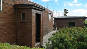A wooden cabin with a small window and an open door, surrounded by a gravel path and dense greenery.