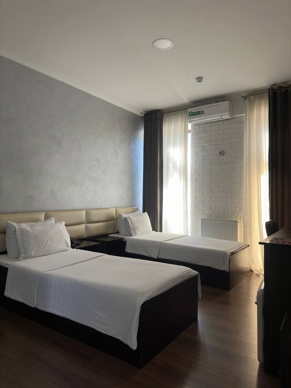 Habitación de hotel con dos camas, una pared de ladrillos blancos, una lámpara en el techo y una ventana con cortinas.