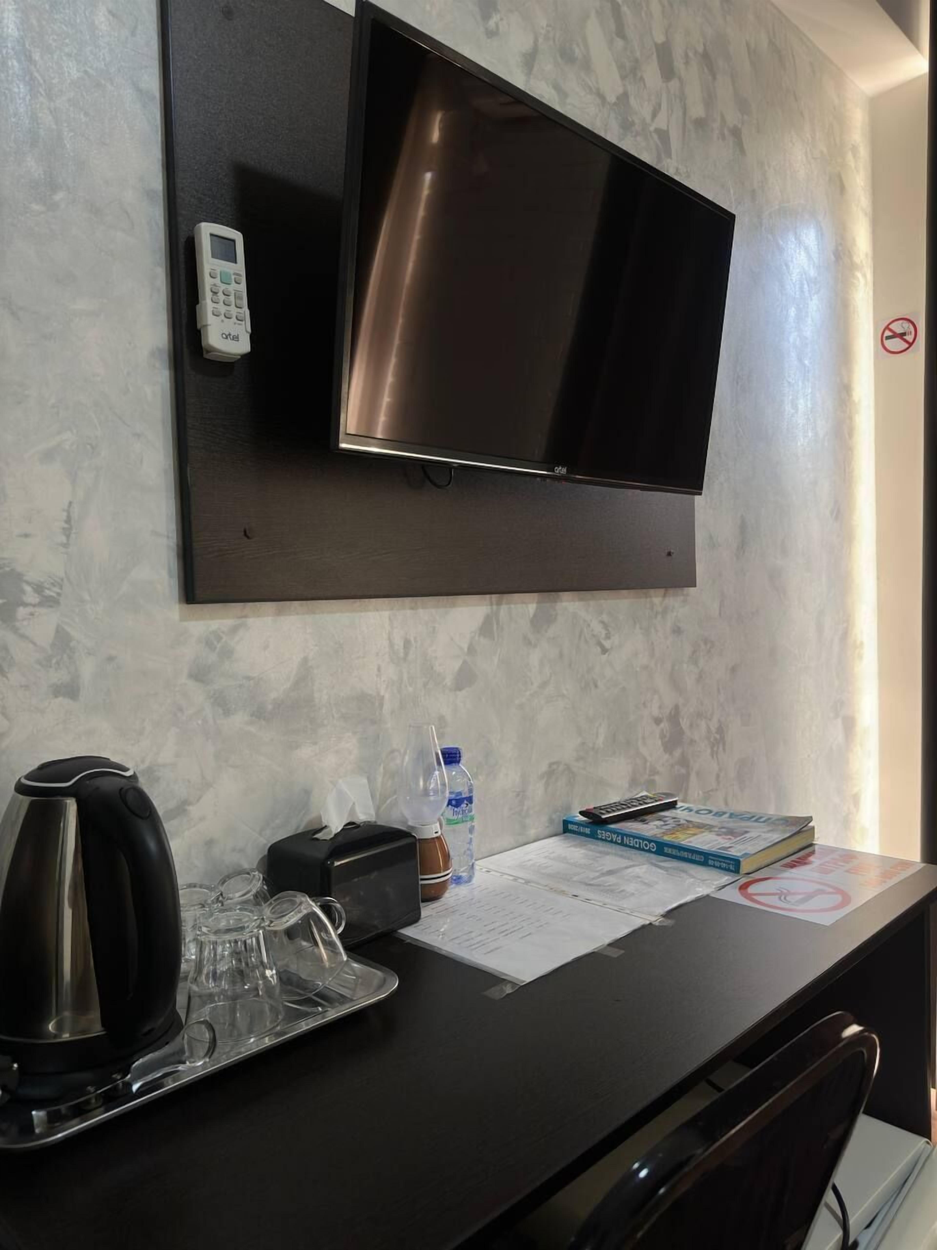 A hotel room with a flat-screen TV mounted on the wall, a desk with a kettle, a bottle of water, a book, and some papers.