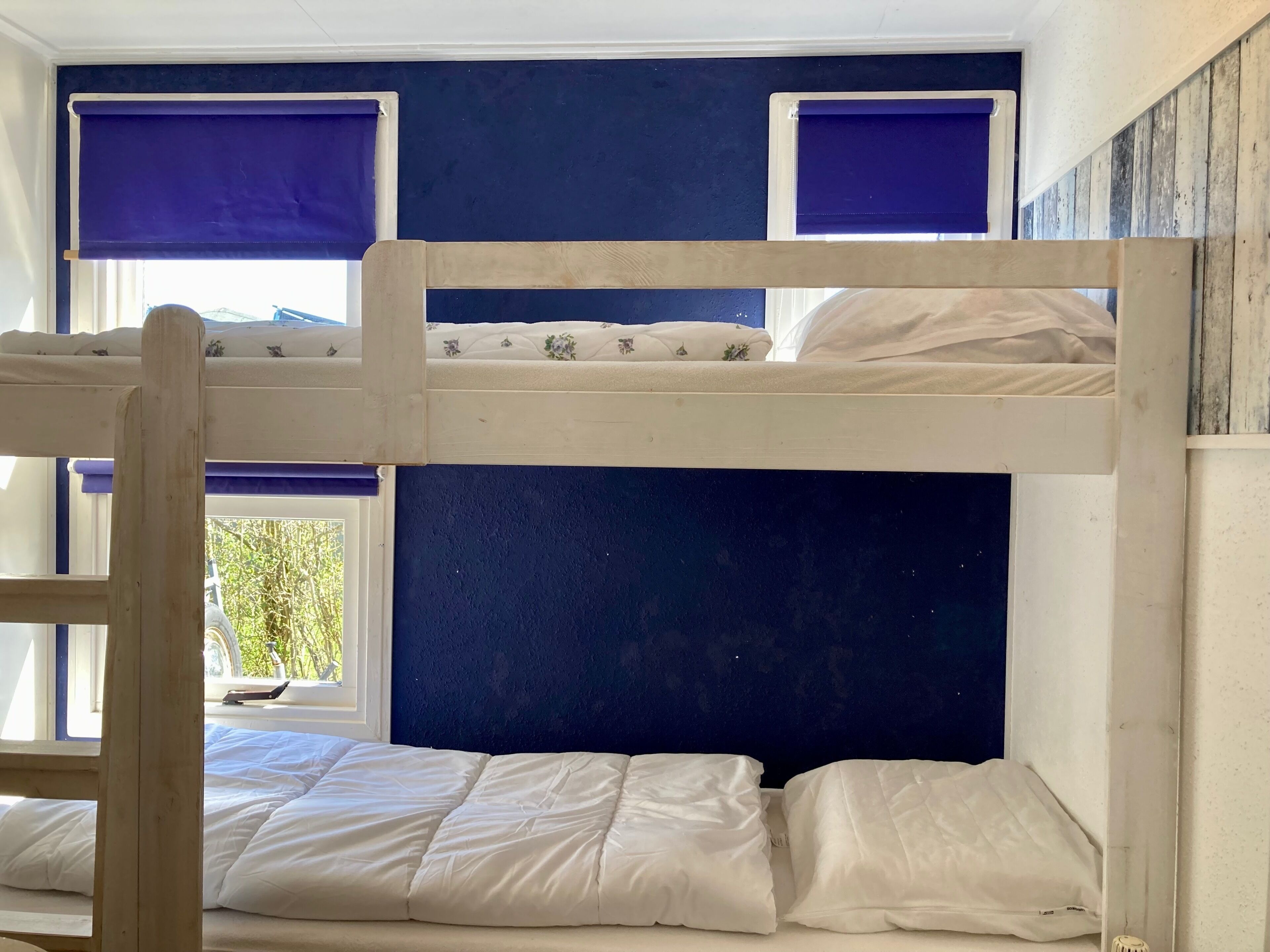 A bunk bed with a headboard against a dark wall, a window with a view of greenery, and a wooden chair in the foreground.