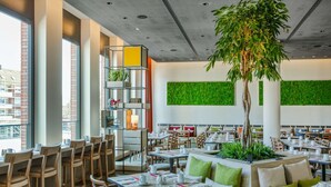 Een modern restaurant met hoge tafels, houten stoelen en een centrale plantenbak met groen.