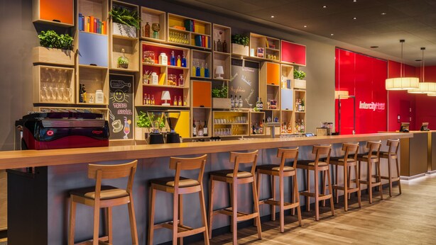 Un espace bar avec des tabourets de bar en bois, des étagères remplies de bouteilles et de verres, et un mur rouge orné du nom de l’hôtel.