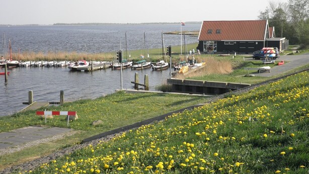 A marina with boats docked, a small building with a red roof, and a grassy area with yellow flowers. - 6 Pers Equipped Holiday Home Behind the Dyke of the Lauwersmeer (Anjum)