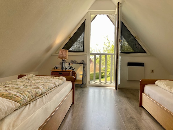 A bedroom with two beds, a window with a view of greenery, a radiator, and a small table with a lamp.