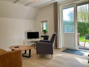 A modern living room with a flat-screen TV, a round wooden table, a grey armchair, and a large sliding glass door leading to an outdoor area. - House Olivia 6p With Private Garden and Close to Lake Lauwersmeer (Anjum)