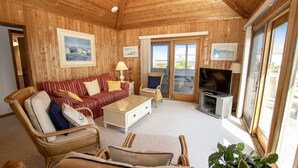Ein Wohnzimmer mit Holzboden, einer Couch, Sesseln, einem Fernseher und einem Balkon mit Glastüren.